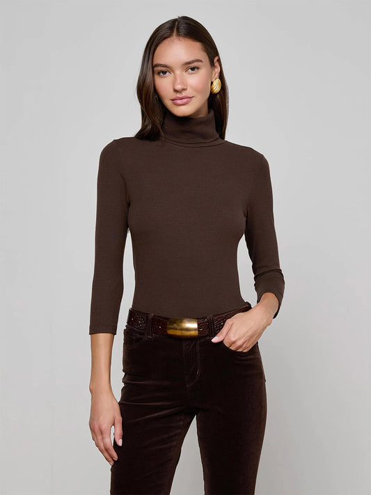 A woman wears the L'Agence Aja Turtleneck in Coffee Bean with dark brown pants and gold earrings, standing against a plain light gray background.
