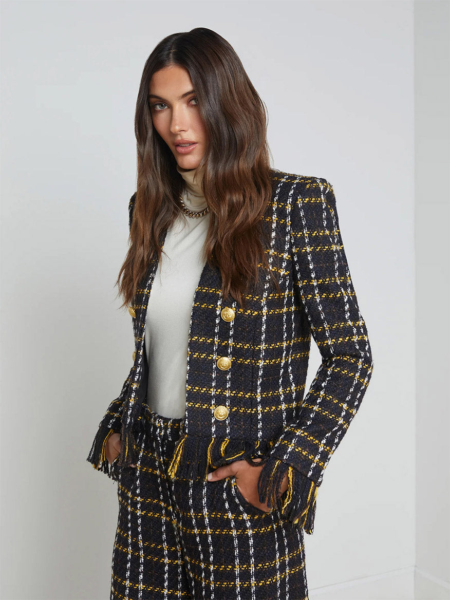 Wearing the L'Agence Belmont Fringe Jacket in Black Yellow Multi Plaid, a person stands against a plain backdrop, exuding a subtle boho vibe with its gold buttons and intricate fringe trim.