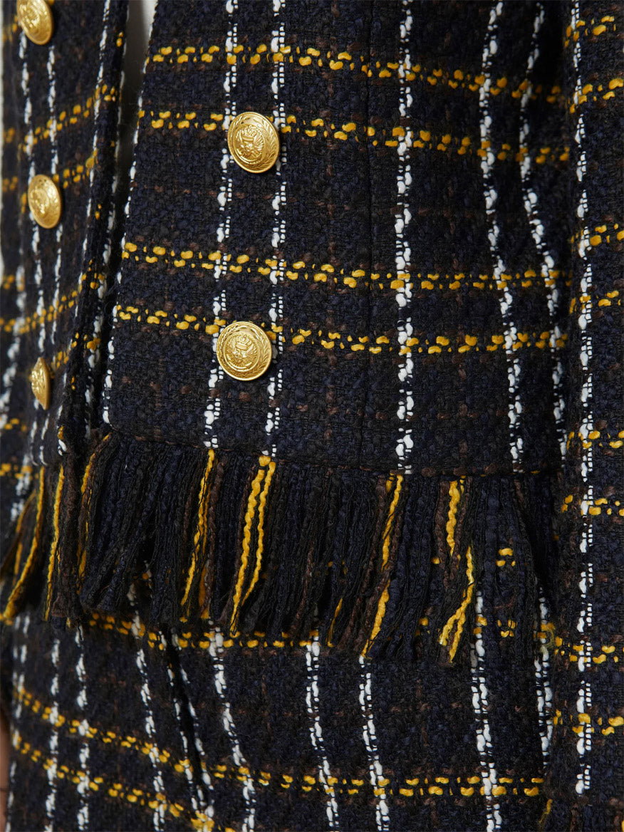 The close-up of the L'Agence Belmont Fringe Jacket in Black Yellow Multi Plaid features a fabric with yellow, white, and dark stripes, complemented by gold buttons and fringe trim detailing.