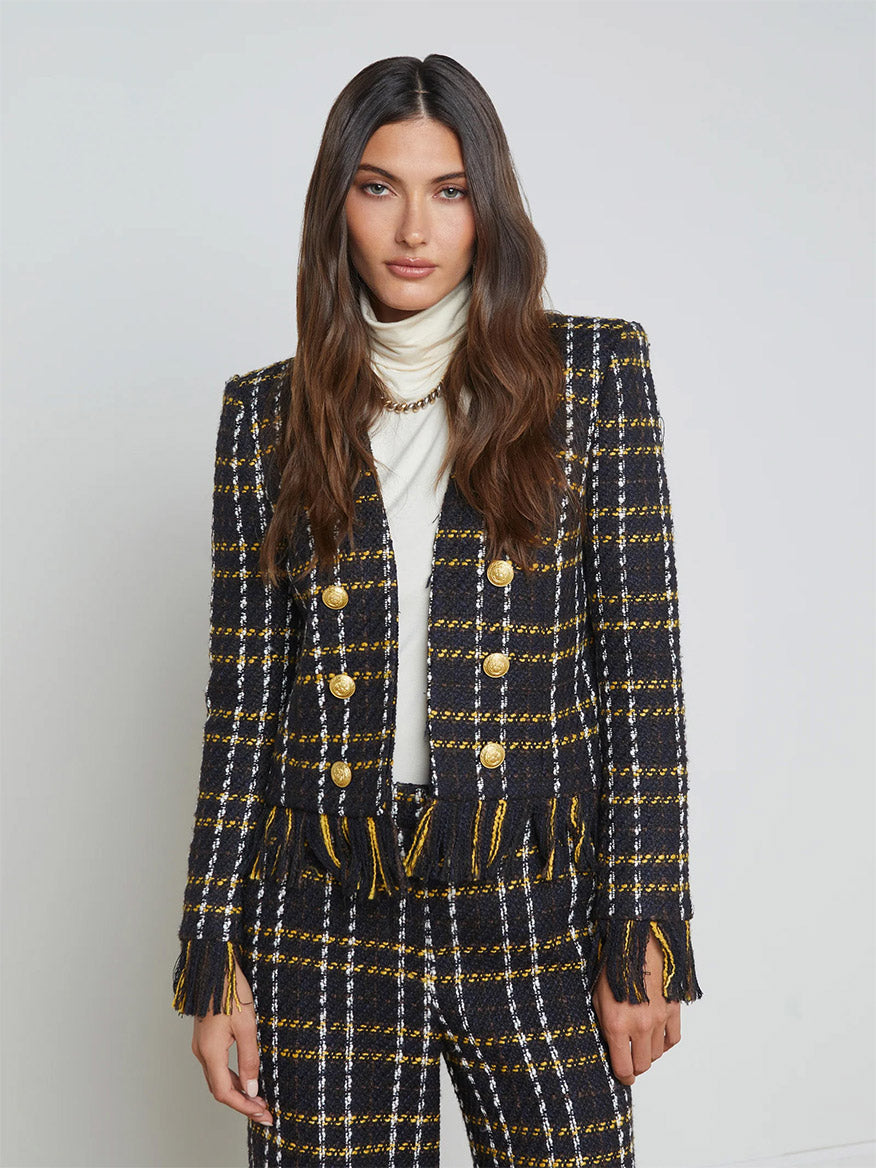 A woman stands in a boho-inspired ensemble, featuring the L'Agence Belmont Fringe Jacket in Black Yellow Multi Plaid, adorned with gold buttons and fringe trim, complemented by a white turtleneck against a plain background.