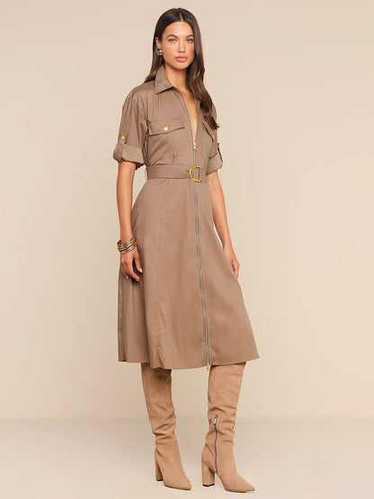 A woman wears the L’Agence Callen Shirt Dress in Cardamom—a tan collared shirt dress crafted from a cotton-blend poplin with an exposed front zip, short sleeves, and a coordinating belt—paired with tan knee-high heeled boots as she stands against a light beige background.