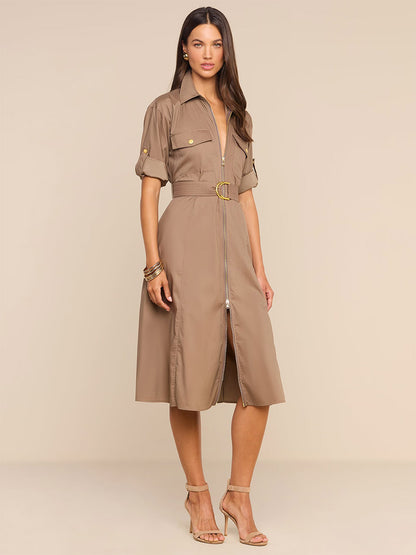 A woman wearing the L’Agence Callen Shirt Dress in Cardamom—a khaki cotton-blend poplin dress featuring an exposed front zip, short sleeves, and gold buttons—paired with beige heeled sandals, stands against a plain beige background.