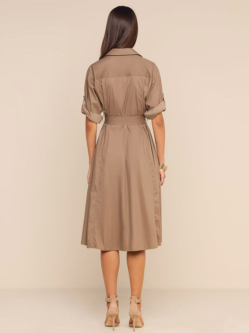 A woman stands with her back to the camera, wearing the L’Agence Callen Shirt Dress in Cardamom— a belted, knee-length cotton-blend poplin piece—paired with heeled sandals, set against a plain beige background.