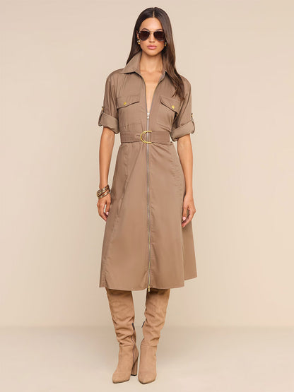 A woman wearing the L’Agence Callen Shirt Dress in Cardamom—featuring rolled sleeves and a belted waist—with sunglasses and knee-high tan boots, stands against a beige background.