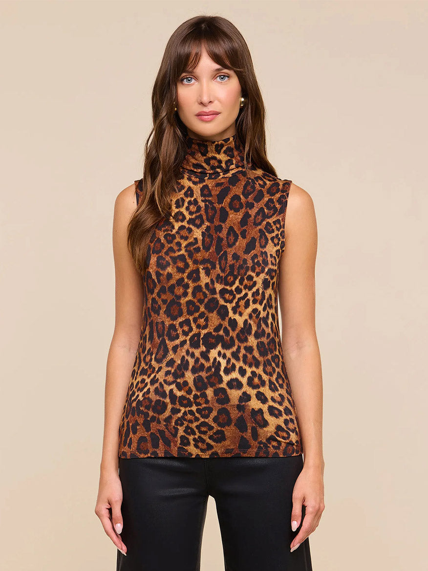 A woman with straight brown hair and a neutral expression stands against a plain background, wearing the L’Agence Ceci Sleeveless Turtleneck in Tan Multi Javan Leopard and black pants.