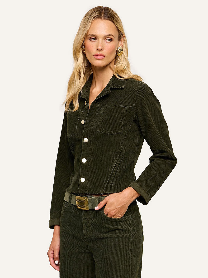 A woman wearing the L’Agence Janelle Corduroy Jacket in Army and matching pants, paired with a belt and gold earrings, stands against a plain light background.