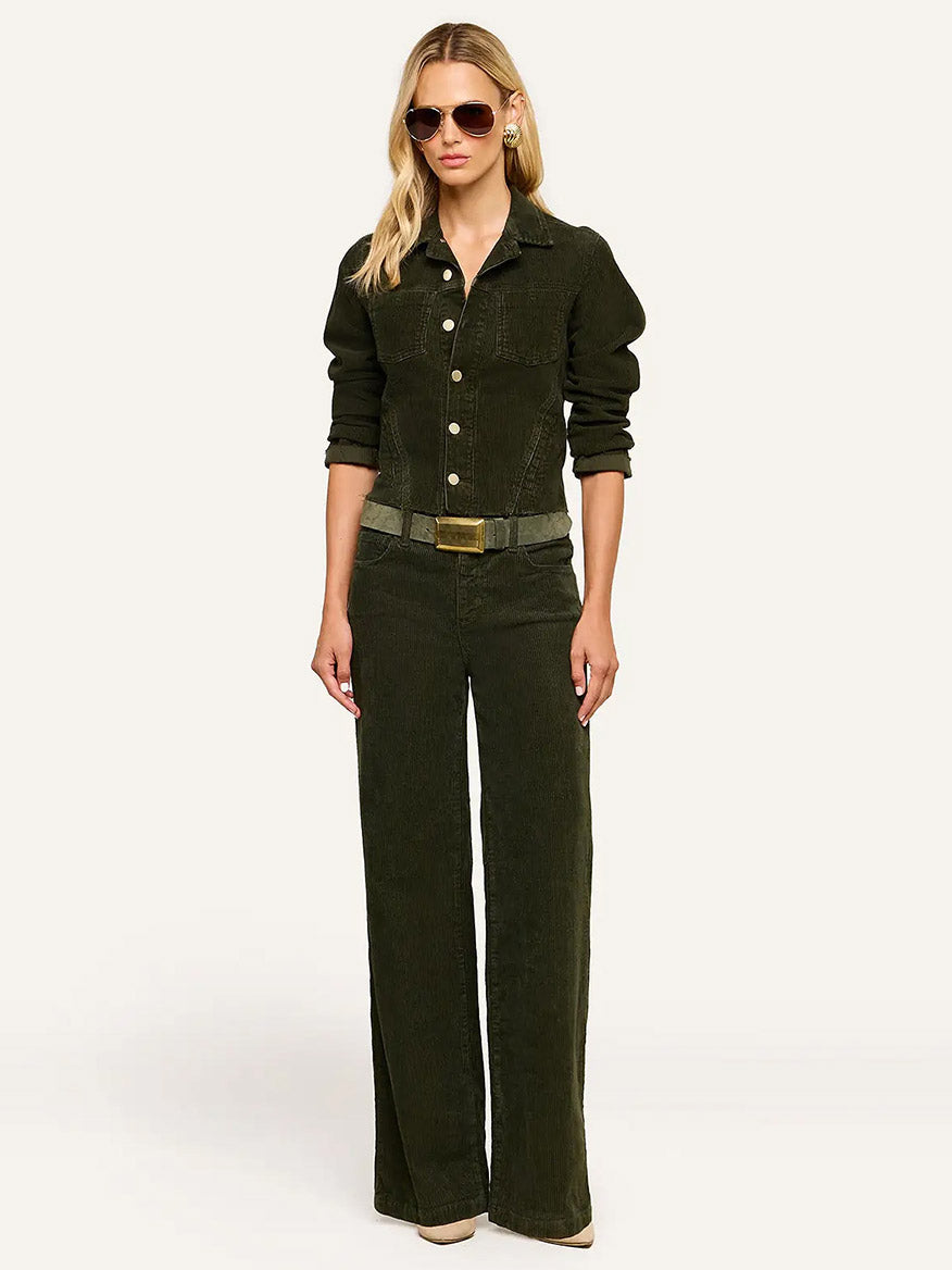 A woman stands against a plain background, wearing the L’Agence Janelle Corduroy Jacket in Army layered over an olive green stretch corduroy jumpsuit with wide-leg pants and a belt, styled with sunglasses and beige heels.