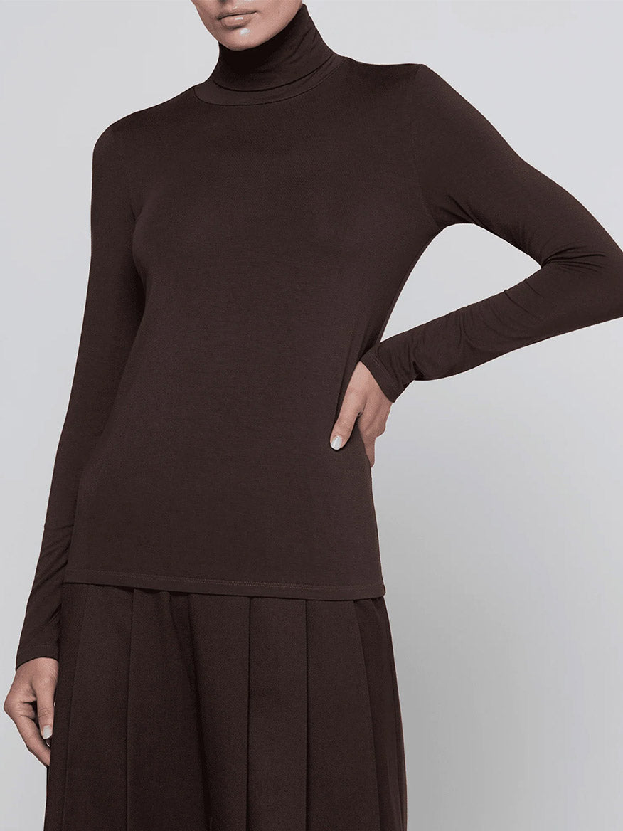 Wearing the L’Agence Lani Turtleneck in Dark Java with a matching pleated skirt, the person poses with one hand on their hip against a plain background.