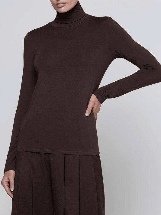 Wearing the L’Agence Lani Turtleneck in Dark Java with a matching pleated skirt, the person poses with one hand on their hip against a plain background.