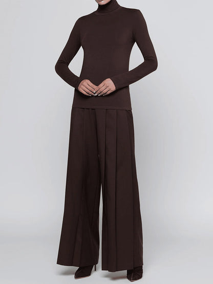 A person wearing the L’Agence Lani Turtleneck in Dark Java and wide-legged pleated brown pants, standing against a plain light background.