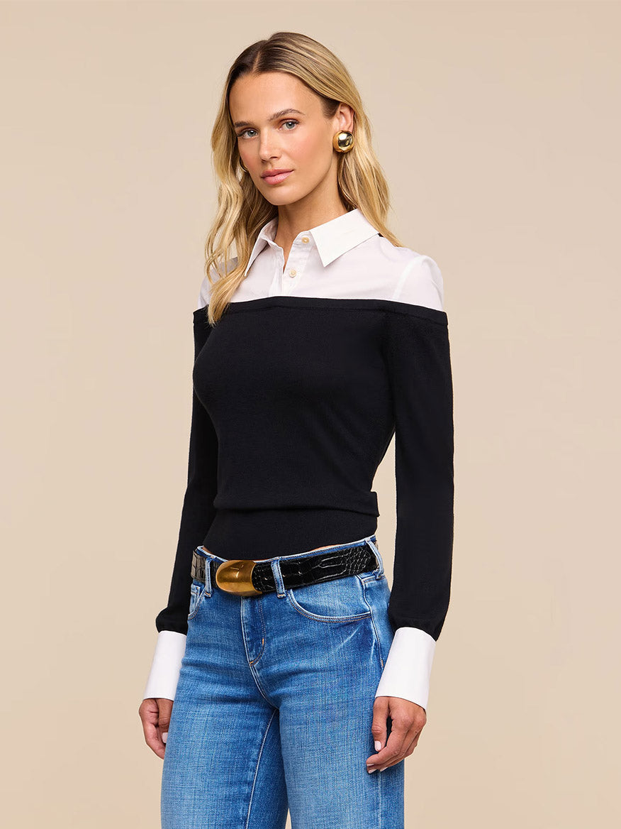 A woman stands against a beige background wearing the L’Agence Mathilde Poplin Collar Pullover in Black/White styled with blue jeans, a black belt with a gold buckle, and gold earrings.