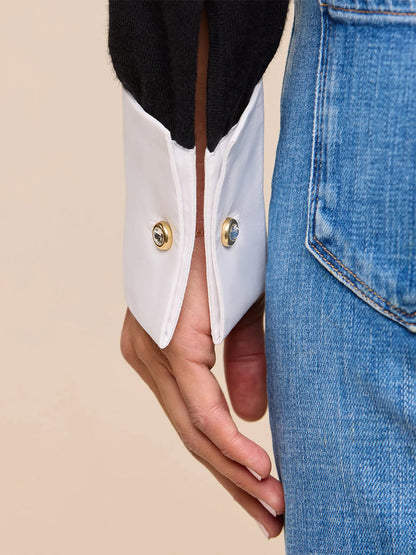 A close-up of a person’s hand resting by their side, wearing blue jeans and the L’Agence Mathilde Poplin Collar Pullover in Black/White with white French cuffs featuring gold buttons.