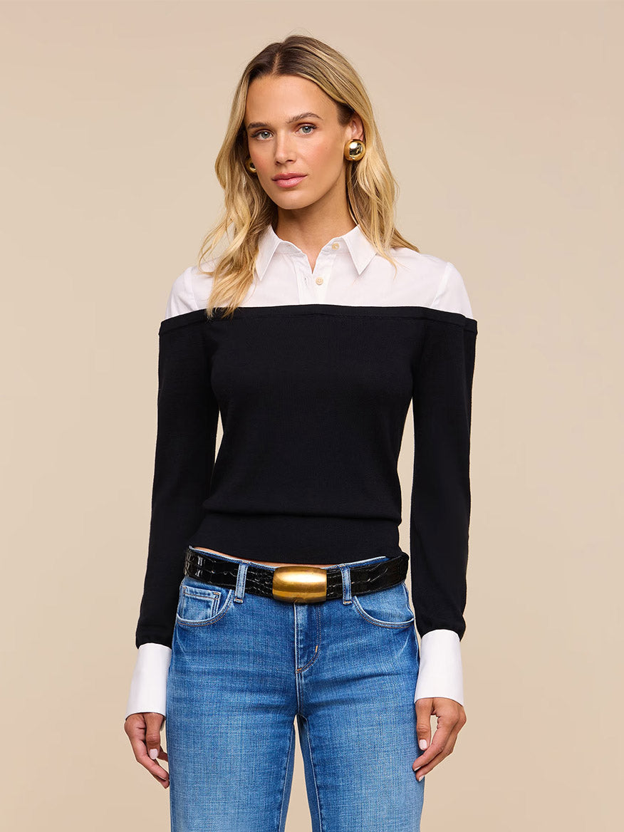 A woman stands facing forward against a beige background, wearing the L’Agence Mathilde Poplin Collar Pullover in Black/White layered over a crisp poplin shirt with French cuffs, paired with blue jeans, a black belt featuring a gold buckle, and gold earrings.