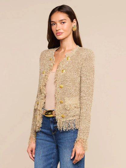 Wearing the L’Agence Mireille Fringe Cardigan in Natural—woven with subtle metallic threads and accented by gold buttons—a woman pairs this textured beige jacket with a tan top, blue jeans, and gold earrings against a neutral background, offering an effortless nod to Fall 2026 fashion.