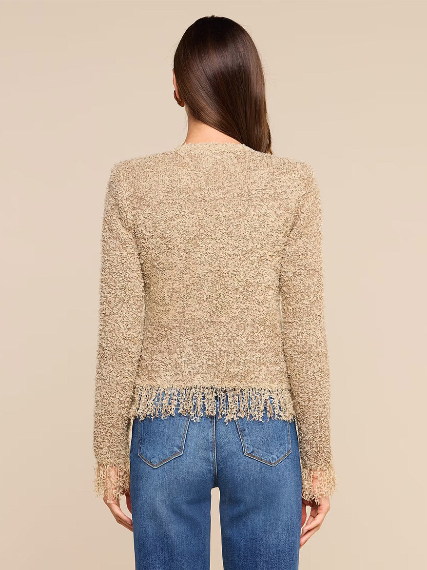 Facing away, a woman wears the L’Agence Mireille Fringe Cardigan in Natural—a textured beige knit subtly accented by metallic threads, with fringe detailing at the hem and cuffs—paired effortlessly with blue jeans against a plain background for a chic Fall 2026 look.