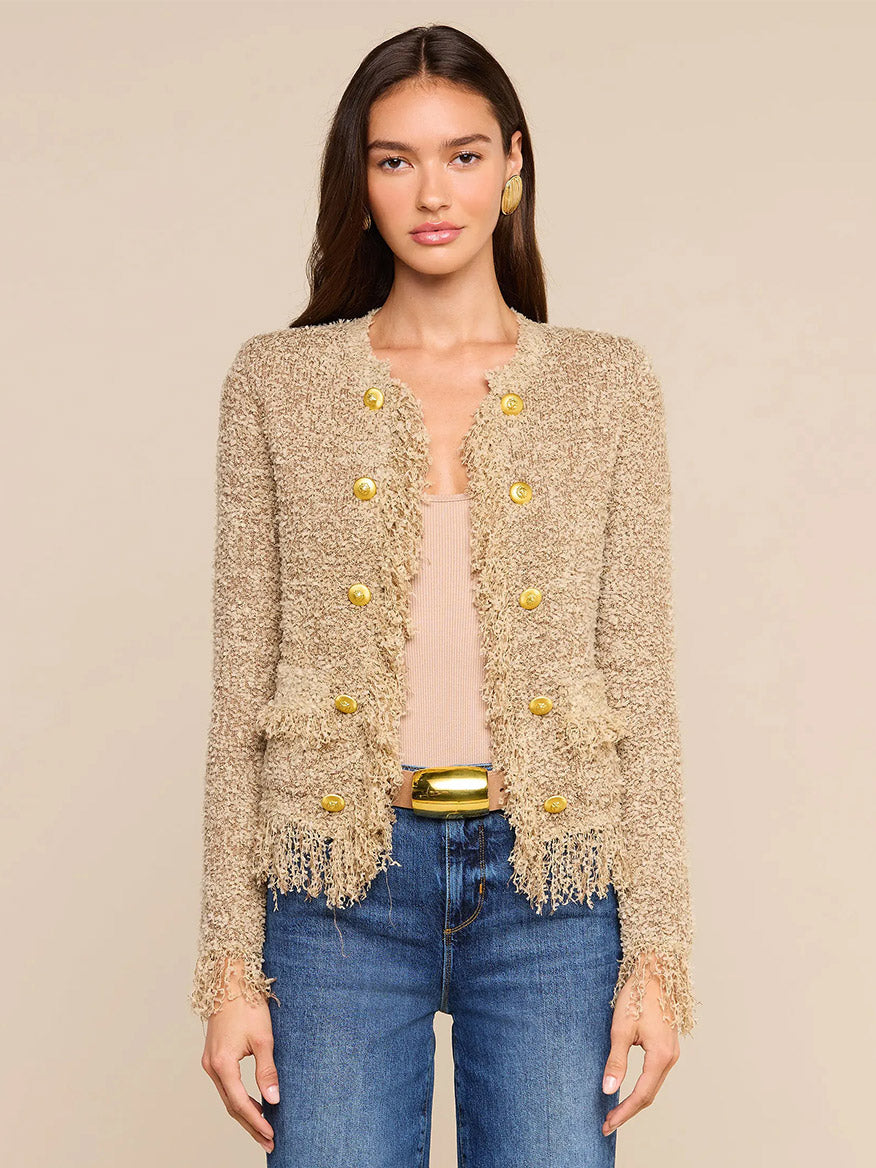 A woman wears the L’Agence Mireille Fringe Cardigan in Natural—woven with subtle metallic threads and finished with gold buttons—over a tan top and blue jeans, standing against a neutral background. This timeless look draws inspiration from Fall 2026 fashion.