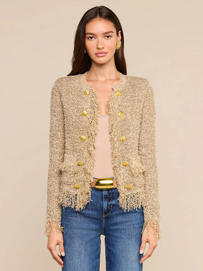 A woman wears the L’Agence Mireille Fringe Cardigan in Natural—woven with subtle metallic threads and finished with gold buttons—over a tan top and blue jeans, standing against a neutral background. This timeless look draws inspiration from Fall 2026 fashion.