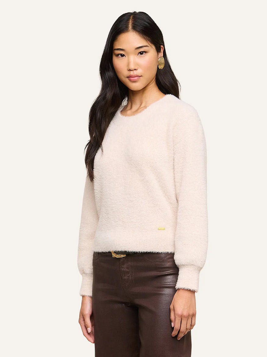A woman with long dark hair wears the L’Agence Nimbus Pullover in Vintage White—a cloud-soft, relaxed fit sweater—paired with brown pants and gold earrings, standing against a plain light background.