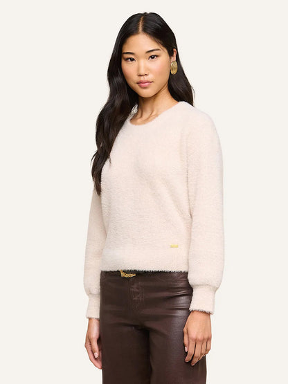 A woman with long dark hair wears the L’Agence Nimbus Pullover in Vintage White—a cloud-soft, relaxed fit sweater—paired with brown pants and gold earrings, standing against a plain light background.