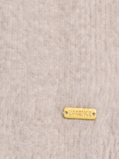 A small gold metal tag engraved with “L’AGENCE” is attached to a beige textured fabric background, evoking the relaxed sophistication and cloud-soft feel of the L’Agence Nimbus Pullover in Vintage White.