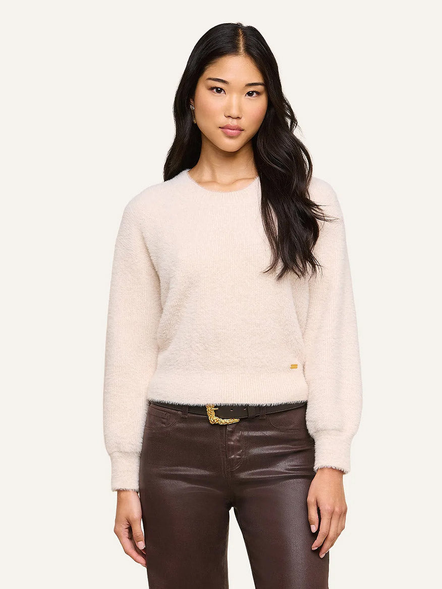 A woman wearing the L’Agence Nimbus Pullover in Vintage White—an ultra-soft, relaxed fit sweater—paired with brown leather pants, stands against a plain light background with a neutral expression, capturing a timeless L’Agence aesthetic.