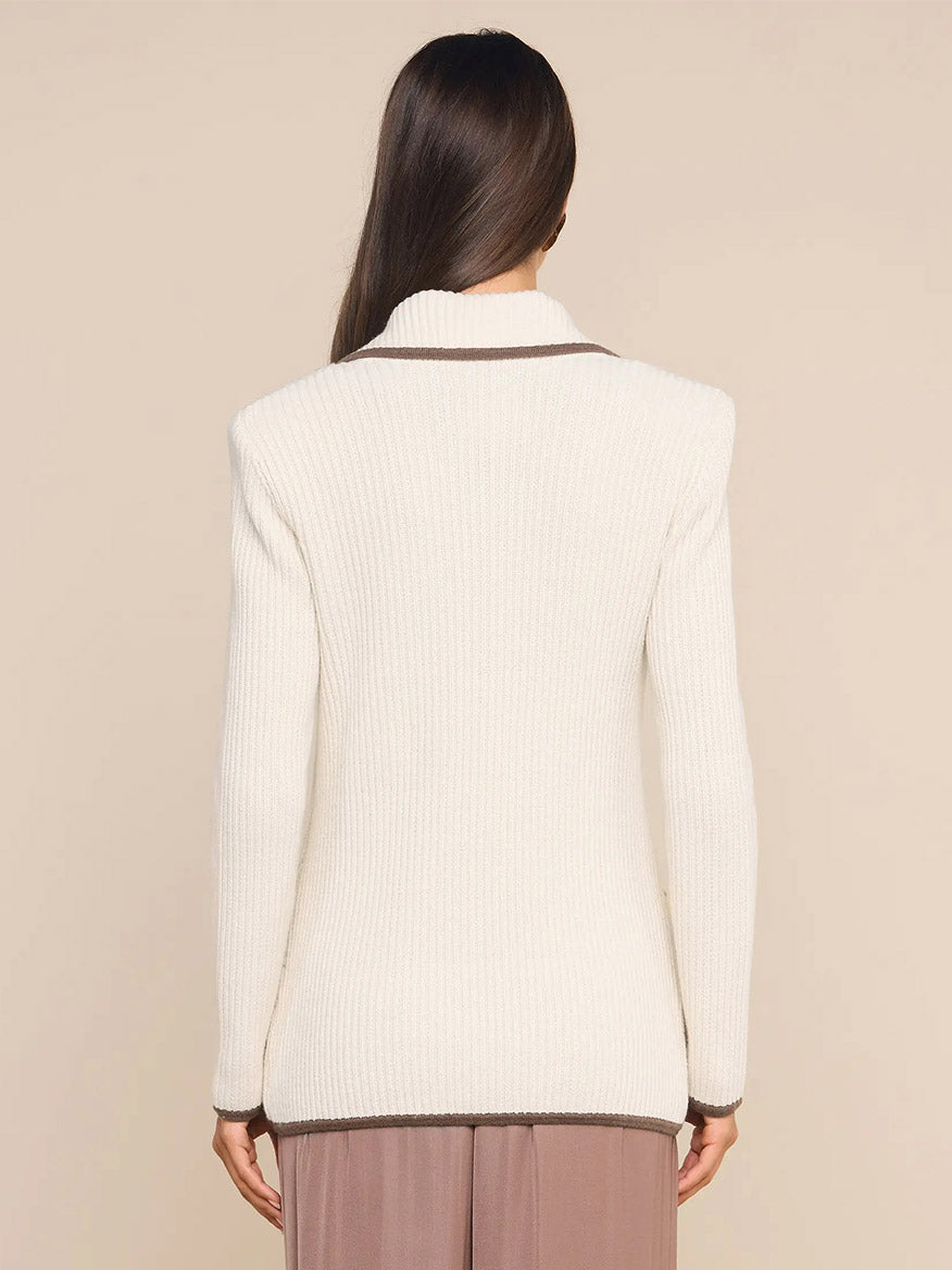 A woman with long brown hair, shown from the back against a neutral background, is wearing the L’Agence Ranch Knit Blazer in Ivory/Dark Cappuccino, featuring a cream-colored ribbed knit design with contrasting brown trim.