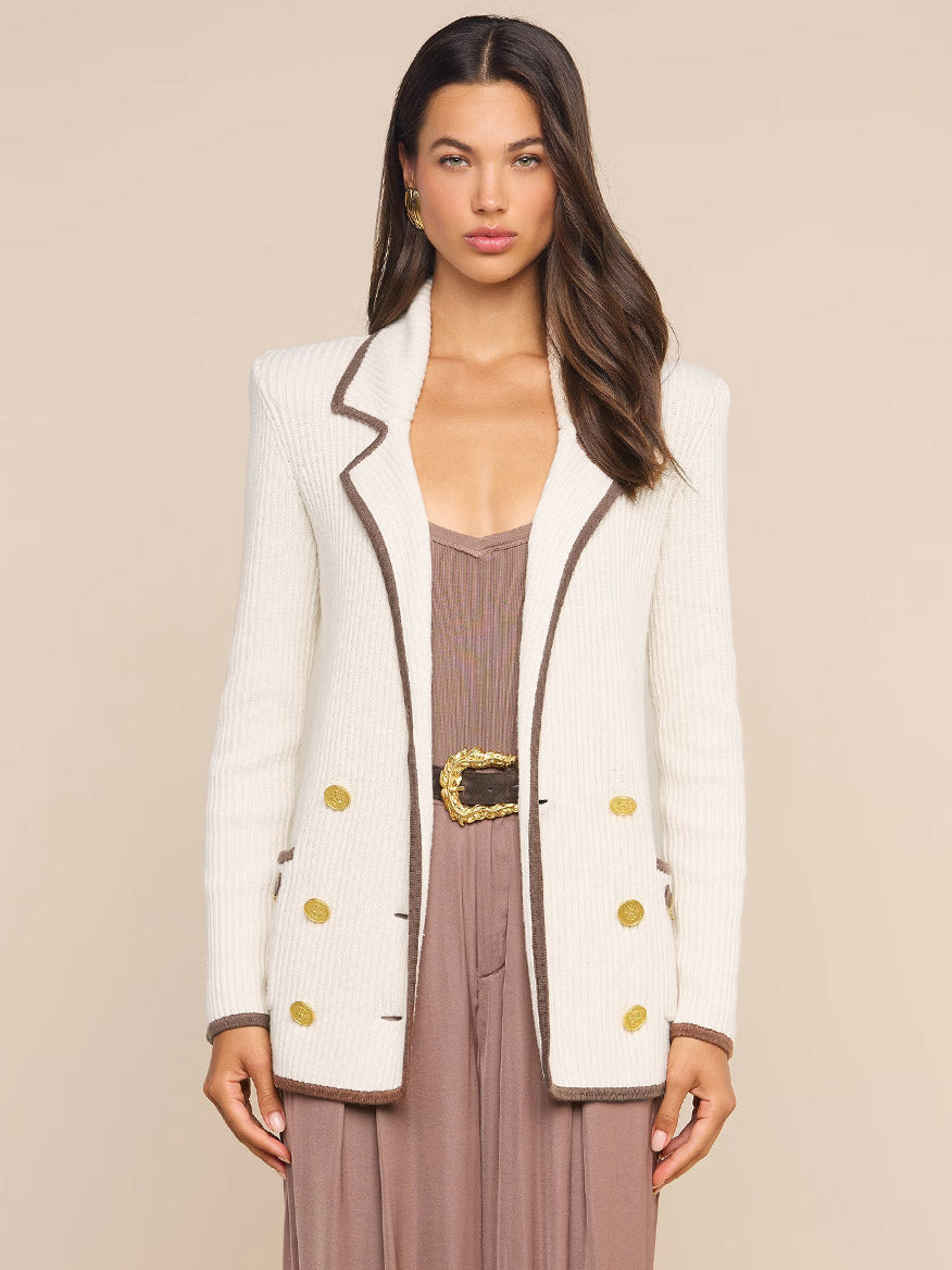 A woman wears the L’Agence Ranch Knit Blazer in Ivory/Dark Cappuccino, featuring brown trim and gold buttons, layered over a mauve dress and standing against a beige background.