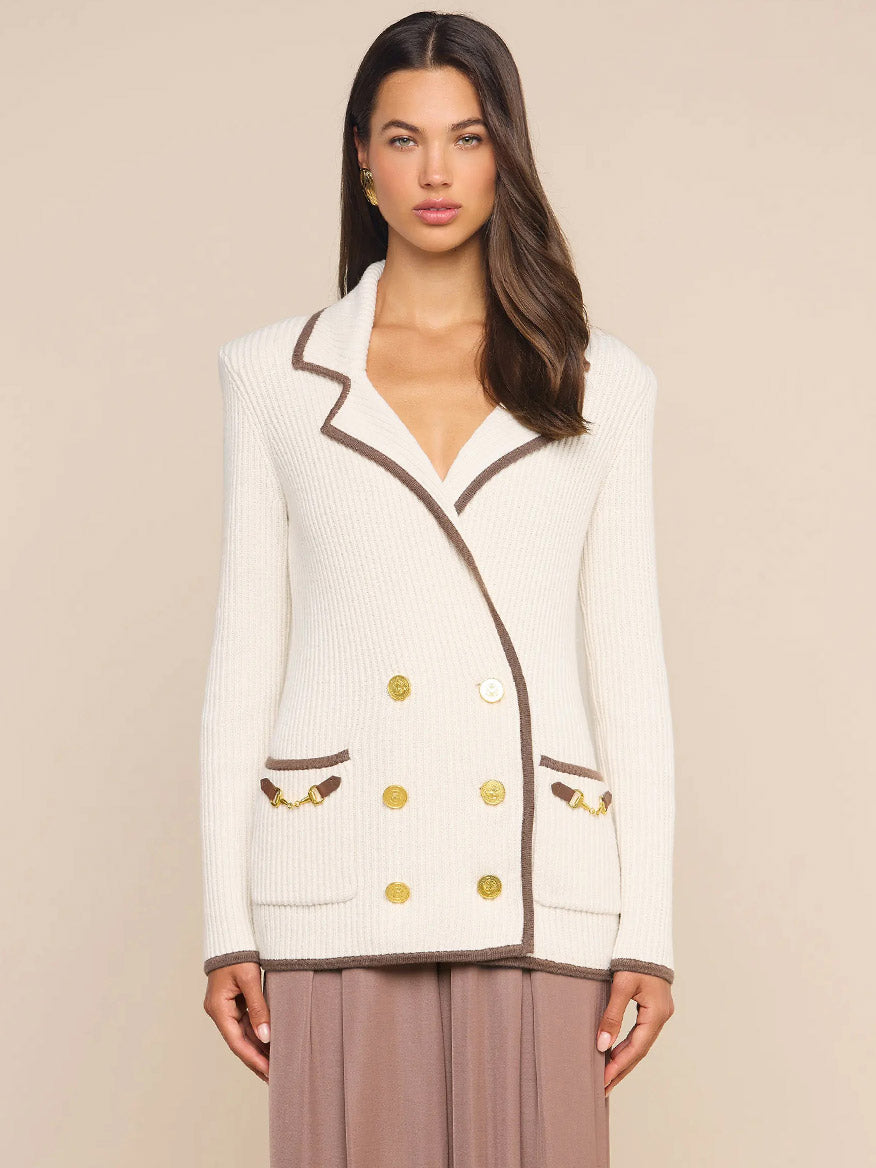 A woman stands against a plain beige background wearing the L’Agence Ranch Knit Blazer in Ivory/Dark Cappuccino, a cream double-breasted ribbed knit blazer detailed with brown trim, gold buttons, and front pockets.