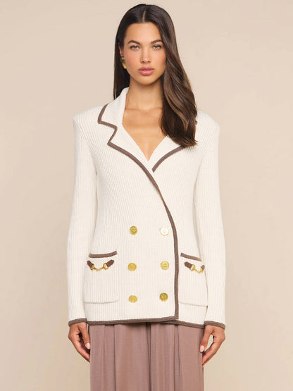 A woman stands against a plain beige background wearing the L’Agence Ranch Knit Blazer in Ivory/Dark Cappuccino, a cream double-breasted ribbed knit blazer detailed with brown trim, gold buttons, and front pockets.