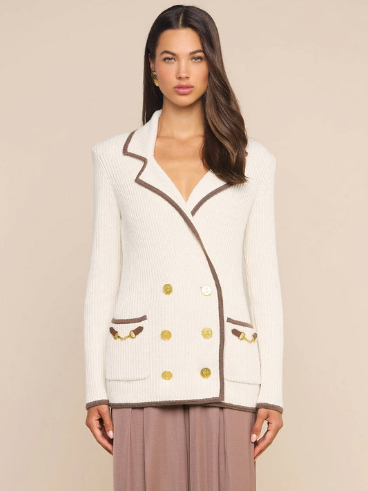A woman stands against a plain beige background wearing the L’Agence Ranch Knit Blazer in Ivory/Dark Cappuccino, a cream double-breasted ribbed knit blazer detailed with brown trim, gold buttons, and front pockets.