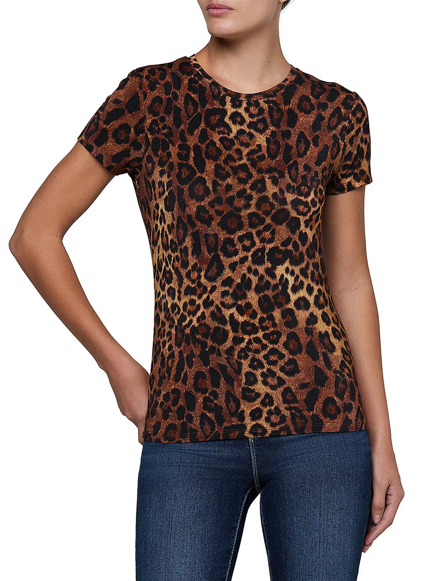 A person stands against a white background with one hand on hip, wearing the L’Agence Ressi Short Sleeve Crew in Tan Multi Javan Leopard—a women’s lightweight jersey tee featuring the signature Ressi silhouette for effortless style, paired with blue jeans.