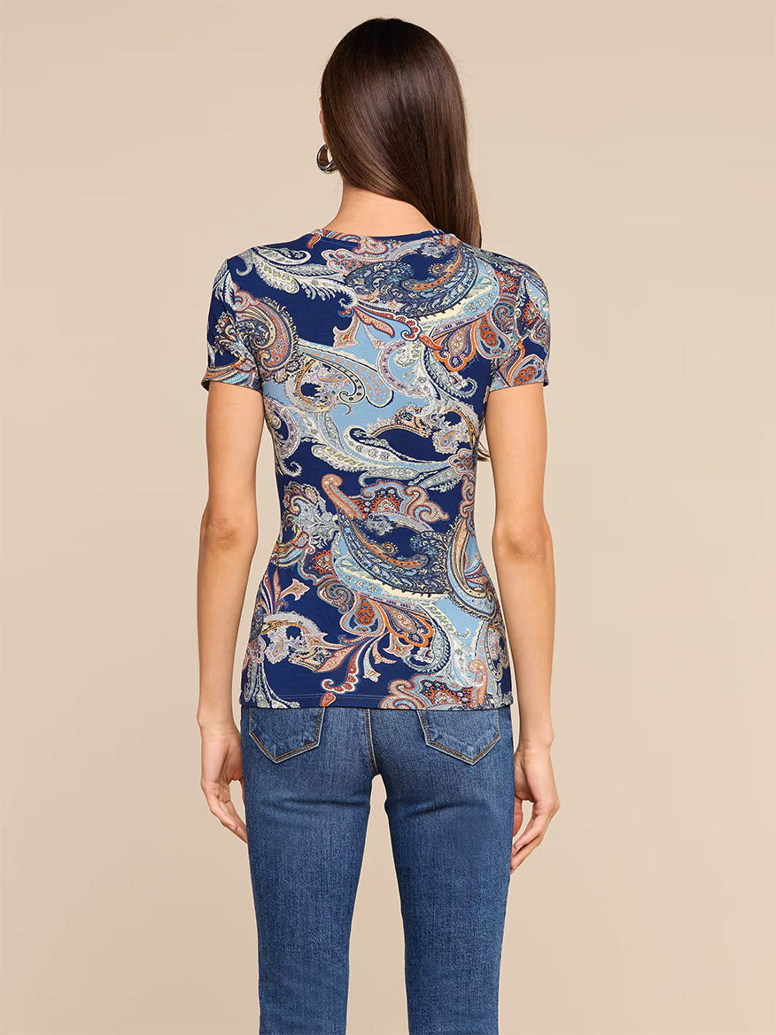 A woman, seen from behind and standing against a plain beige background, wears the L’Agence Ressi Fitted Tee in Midnight Multi Besarabian Paisley paired with blue jeans.