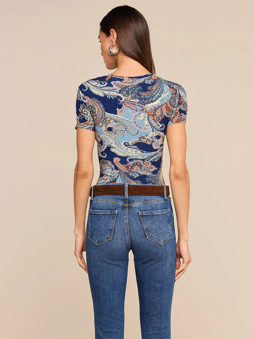 A woman stands with her back to the camera, wearing the L’Agence Ressi Fitted Tee in Midnight Multi Besarabian Paisley, paired with hoop earrings and blue jeans against a plain beige background. This effortlessly sophisticated look is made in Los Angeles.