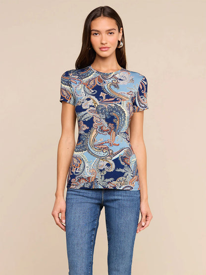 A woman stands against a beige background, wearing the L’Agence Ressi Fitted Tee in Midnight Multi Besarabian Paisley paired with blue jeans. Her long brown hair and hoop earrings complete this effortlessly stylish look, made in Los Angeles.