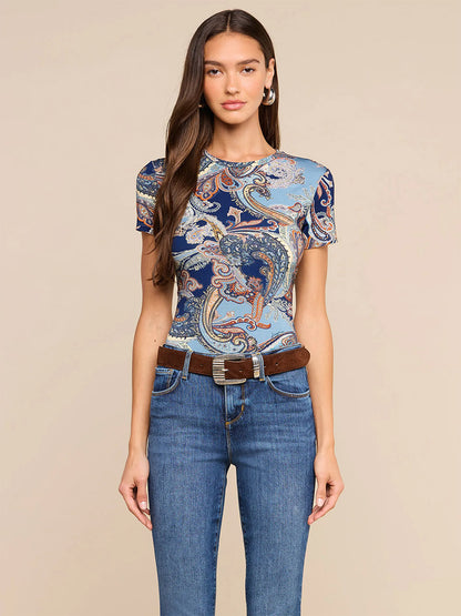 Woman standing against a beige background, wearing the L’Agence Ressi Fitted Tee in Midnight Multi Besarabian Paisley, paired with jeans, a brown belt, and hoop earrings—made in Los Angeles.