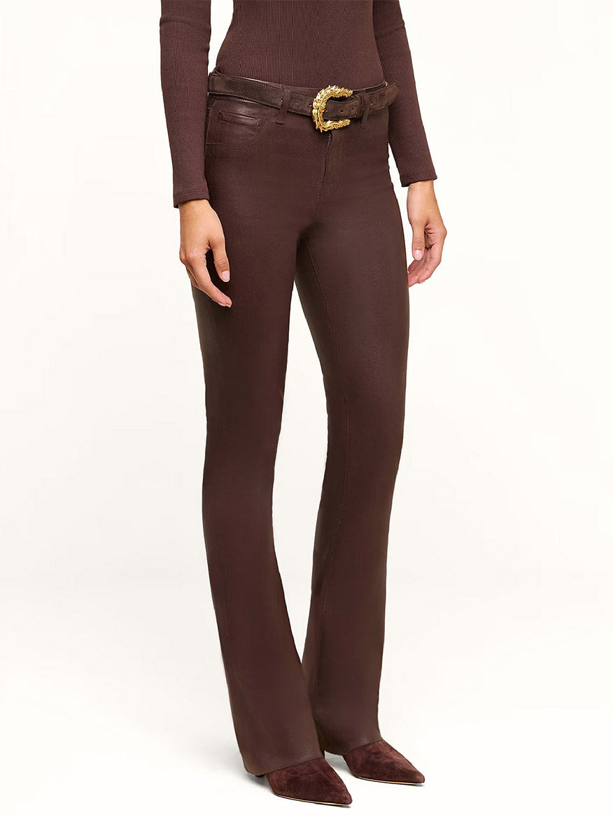 A person wearing the L’Agence Ruth Coated Straight-Leg Jean in Dark Java Coated paired with a matching long-sleeve top, a brown belt with a gold buckle, and brown pointed-toe shoes, standing against a plain background.
