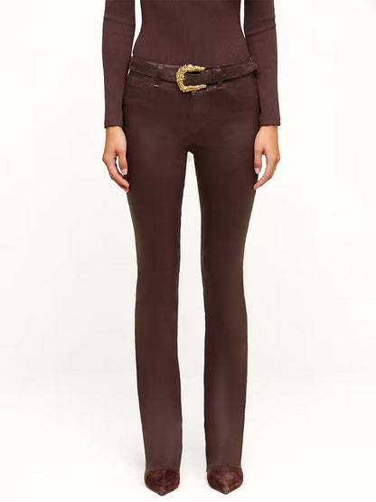 A woman stands against a plain light background wearing a brown long-sleeve top, brown pointed shoes, a brown belt with a gold buckle, and the L’Agence Ruth Coated Straight-Leg Jean in Dark Java Coated.