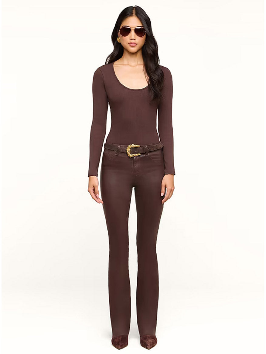 A woman stands against a plain light background, wearing sunglasses, a long-sleeved brown top, and the L’Agence Ruth Coated Straight-Leg Jean in Dark Java Coated with a raw hem, paired with a belt and brown shoes.