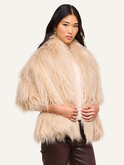 A woman models the L’Agence Sabine Faux Fur Scarf in Biscuit—an elegant, light beige accessory ideal for cool-weather occasions—draped over a pale top and dark pants, standing against a plain light background.