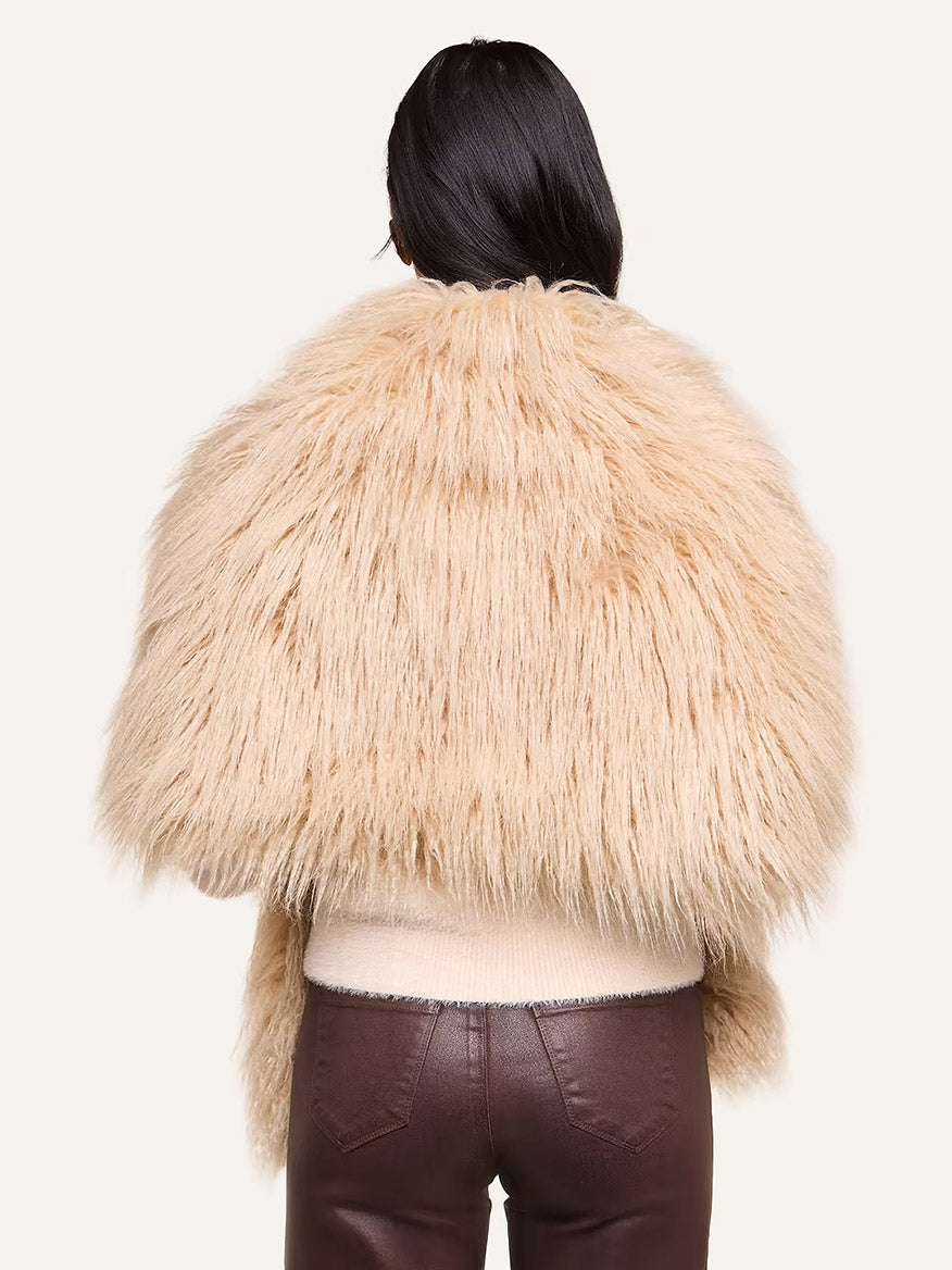 Wearing the L’Agence Sabine Faux Fur Scarf in Biscuit—a chic cool-weather accessory—paired with brown pants, this person is facing away, their long dark hair standing out against a plain light background.