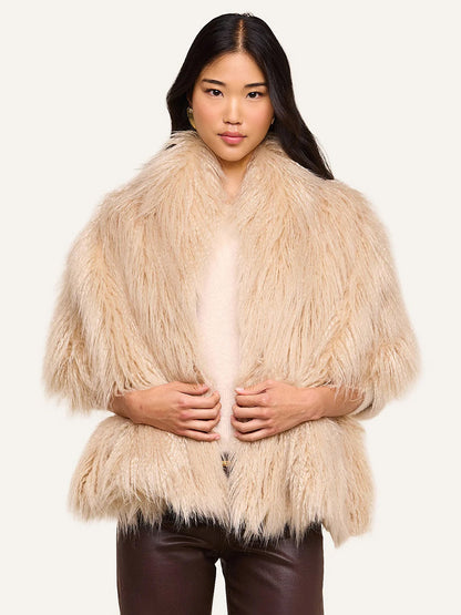 Wearing the L’Agence Sabine Faux Fur Scarf in Biscuit—a plush, light beige accessory perfect for cool weather—draped over a light shirt and dark brown pants, the woman stands against a plain light background.