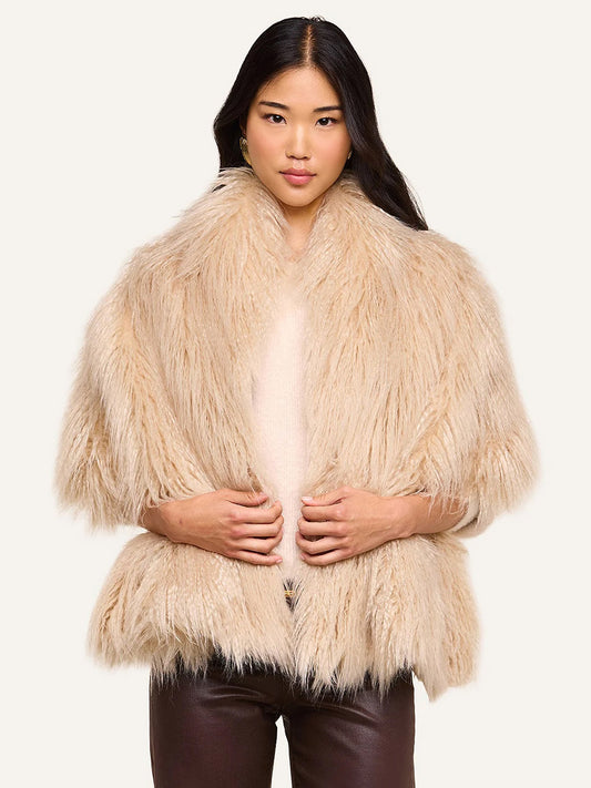 Wearing the L’Agence Sabine Faux Fur Scarf in Biscuit—a plush, light beige accessory perfect for cool weather—draped over a light shirt and dark brown pants, the woman stands against a plain light background.