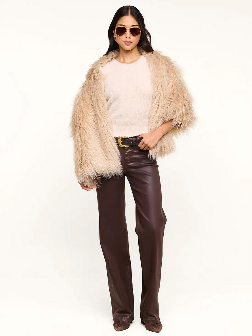 A woman wearing sunglasses drapes the L’Agence Sabine Faux Fur Scarf in Biscuit—a chic cool-weather accessory—over a beige faux fur coat and cream top, paired with brown leather pants and brown shoes, standing against a plain white background. Perfect for a special event or for elevating everyday elegance.