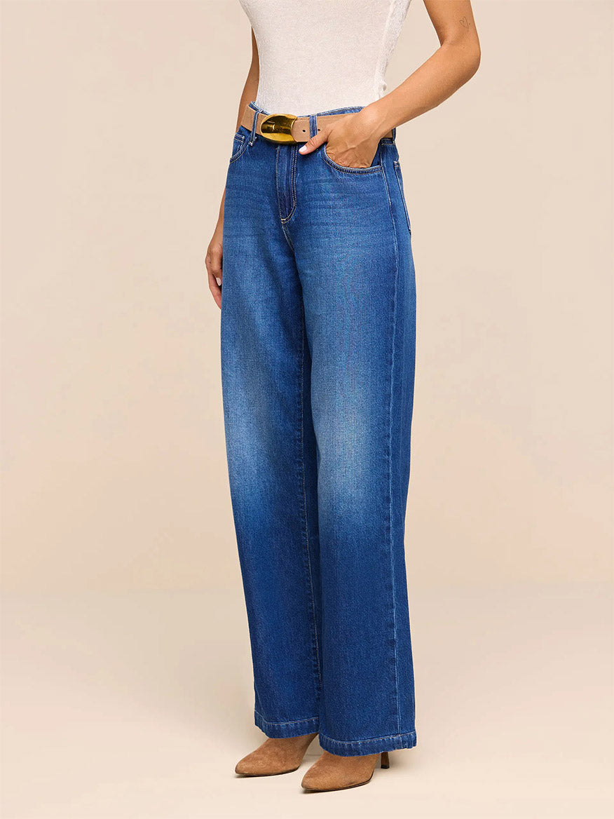 A person wearing the L’Agence Scottie Wide-Leg Jean in Idaho—a high-rise, wide-leg blue jean crafted from nonstretch denim—paired with a white sleeveless top and brown pointed-toe shoes, standing with one hand in their pocket against a plain beige background.