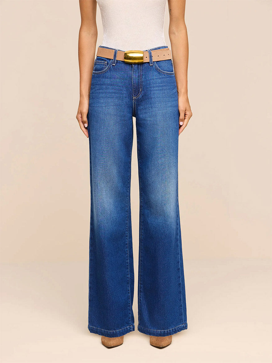 Wearing the L’Agence Scottie Wide-Leg Jean in Idaho—crafted from nonstretch, high-rise blue denim—paired with a light beige belt and a white sleeveless top, this individual stands against a plain light background.