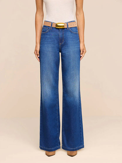 Wearing the L’Agence Scottie Wide-Leg Jean in Idaho—crafted from nonstretch, high-rise blue denim—paired with a light beige belt and a white sleeveless top, this individual stands against a plain light background.