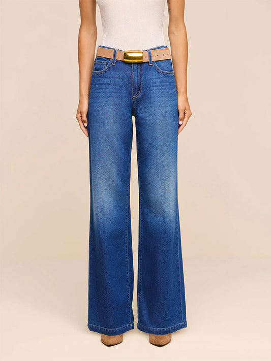 Wearing the L’Agence Scottie Wide-Leg Jean in Idaho—crafted from nonstretch, high-rise blue denim—paired with a light beige belt and a white sleeveless top, this individual stands against a plain light background.