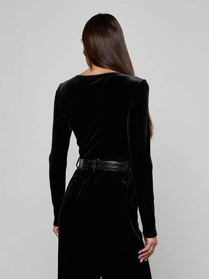 A person with long brown hair faces away, wearing the L'Agence Seraphine Velvet Bodysuit in Black with long sleeves and matching belted pants against a plain light background. This stylish look is made in Los Angeles.