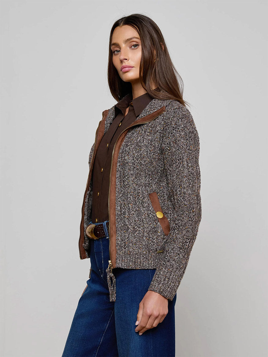 A woman stands against a plain background wearing the L'Agence Shiloh Cable Knit Cardigan in Carafe Brown Multi, a textured button-up piece with brown trim, styled with blue jeans.