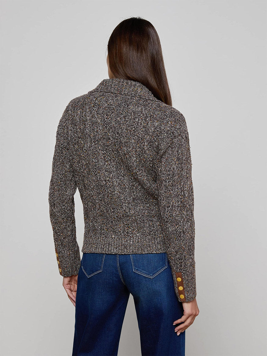 A woman with long brown hair is shown from the back in the L'Agence Shiloh Cable Knit Cardigan in Carafe Brown Multi, featuring gold-buttoned cuffs, styled with blue jeans against a plain background.